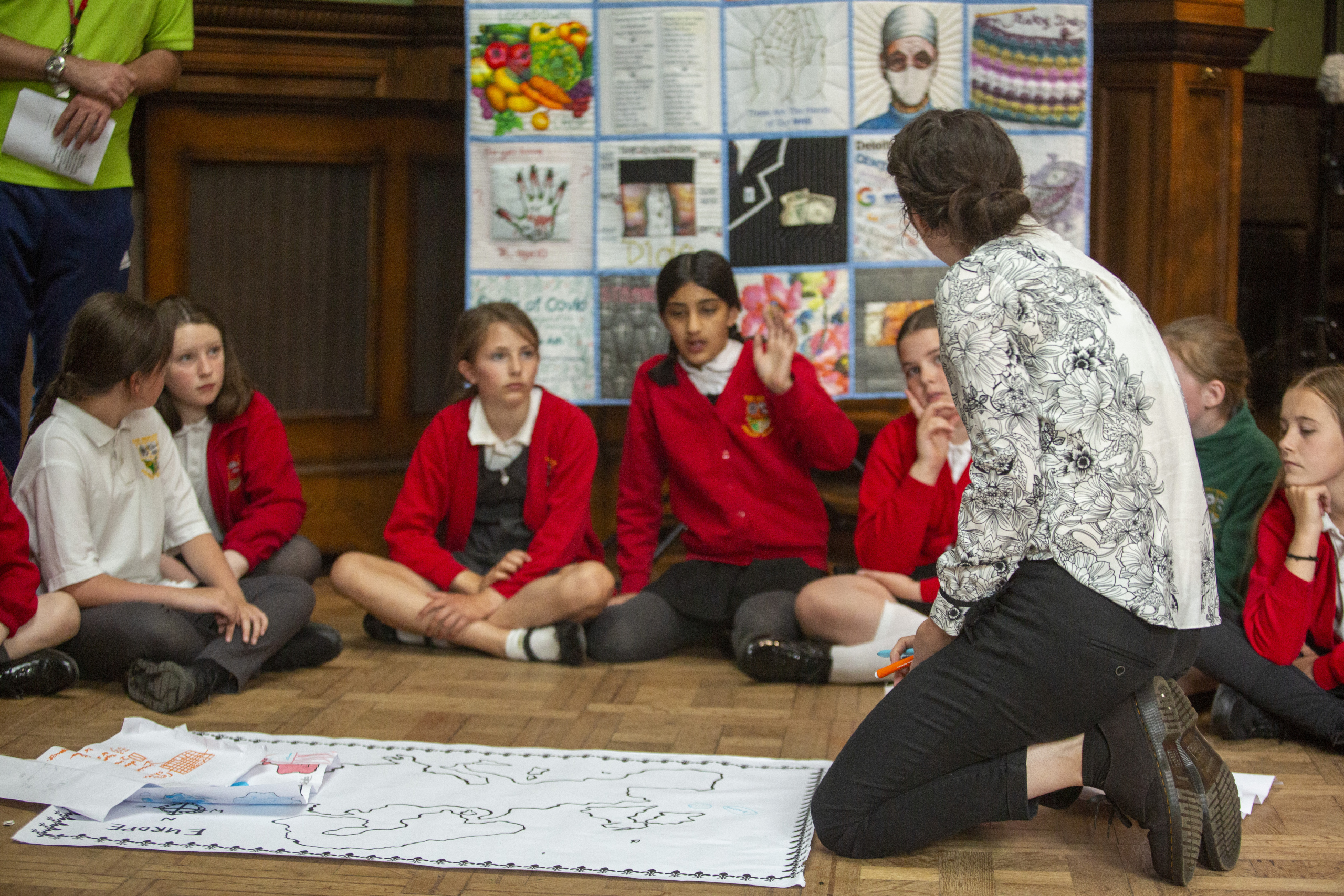 School children in a drama workshop. The children are sat on the floor engaged in a discussion with a facilitator.
