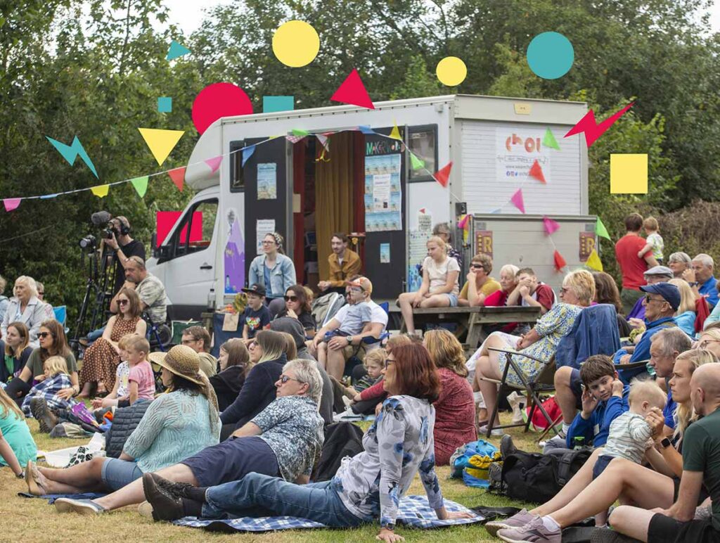 Large crowd sitting on the grass in the summer with bunting, watching a performance