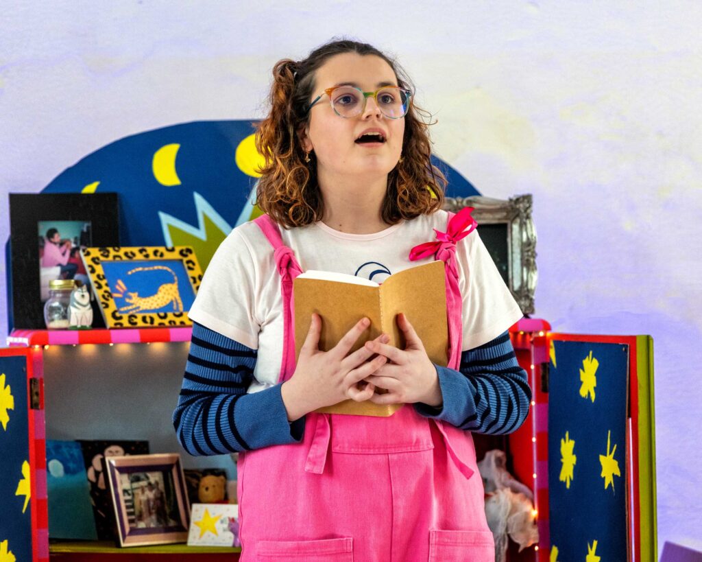 Young person with glasses and pink dungarees singing with an open book
