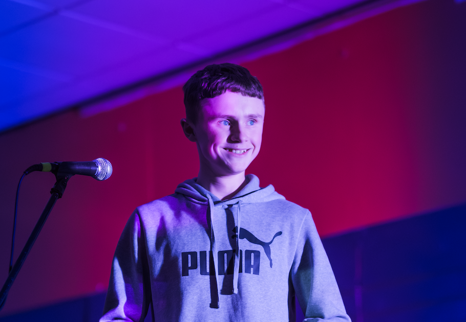 Young person standing at a microphone on stage, holding notes, lit by pink and blue lighting during a live spoken word performance at MakerWorld in Huddersfield.